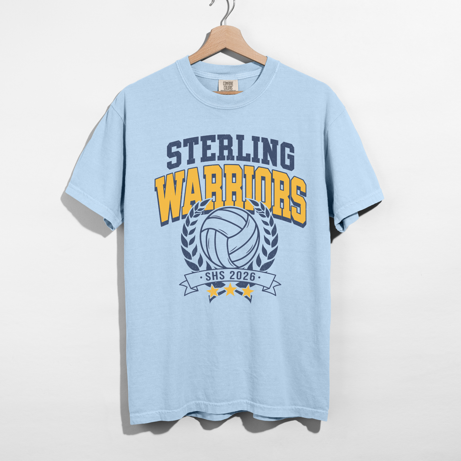 Chambray blue Sterling Warriors Volleyball Comfort Colors graphic tee hanging on wooden hanger featuring navy and gold volleyball school spirit design.