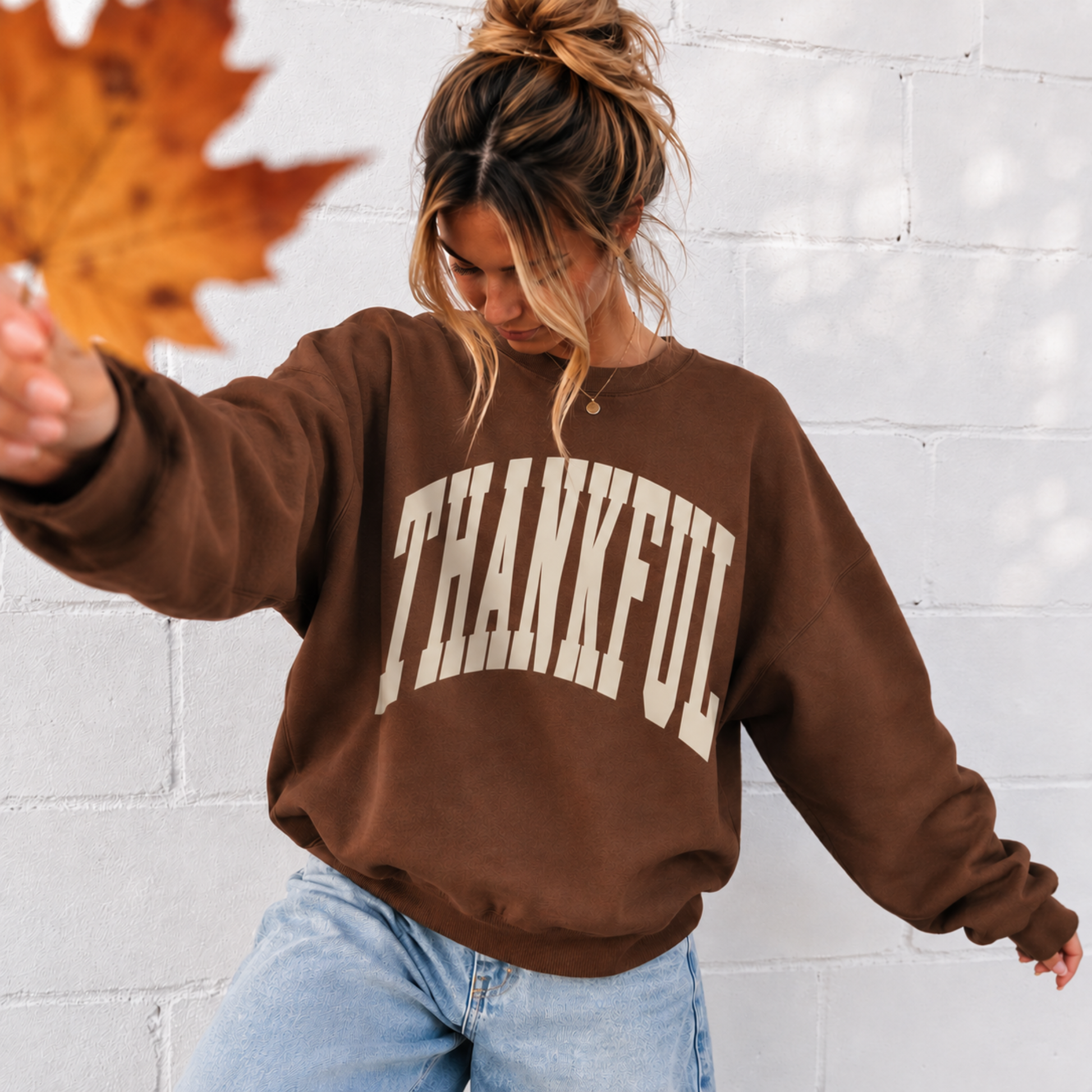 Woman wearing chestnut brown Thankful sweatshirt with cream varsity lettering and jeans for a casual fall outfit