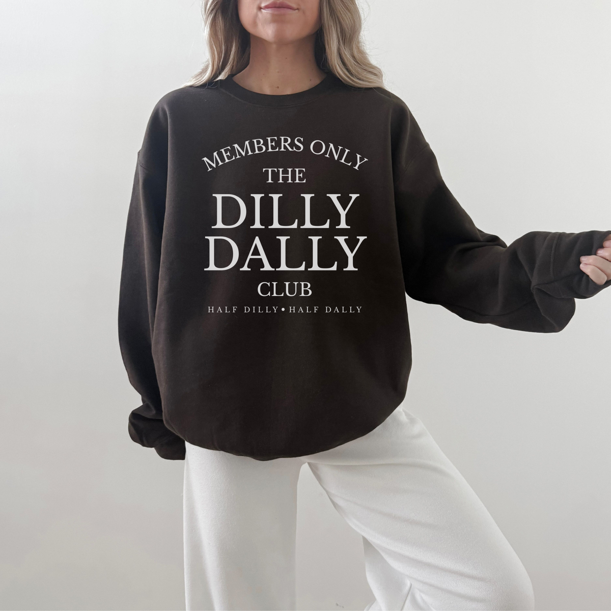 Woman modeling a chocolate brown “Dilly Dally Club” crewneck sweatshirt with white pants.