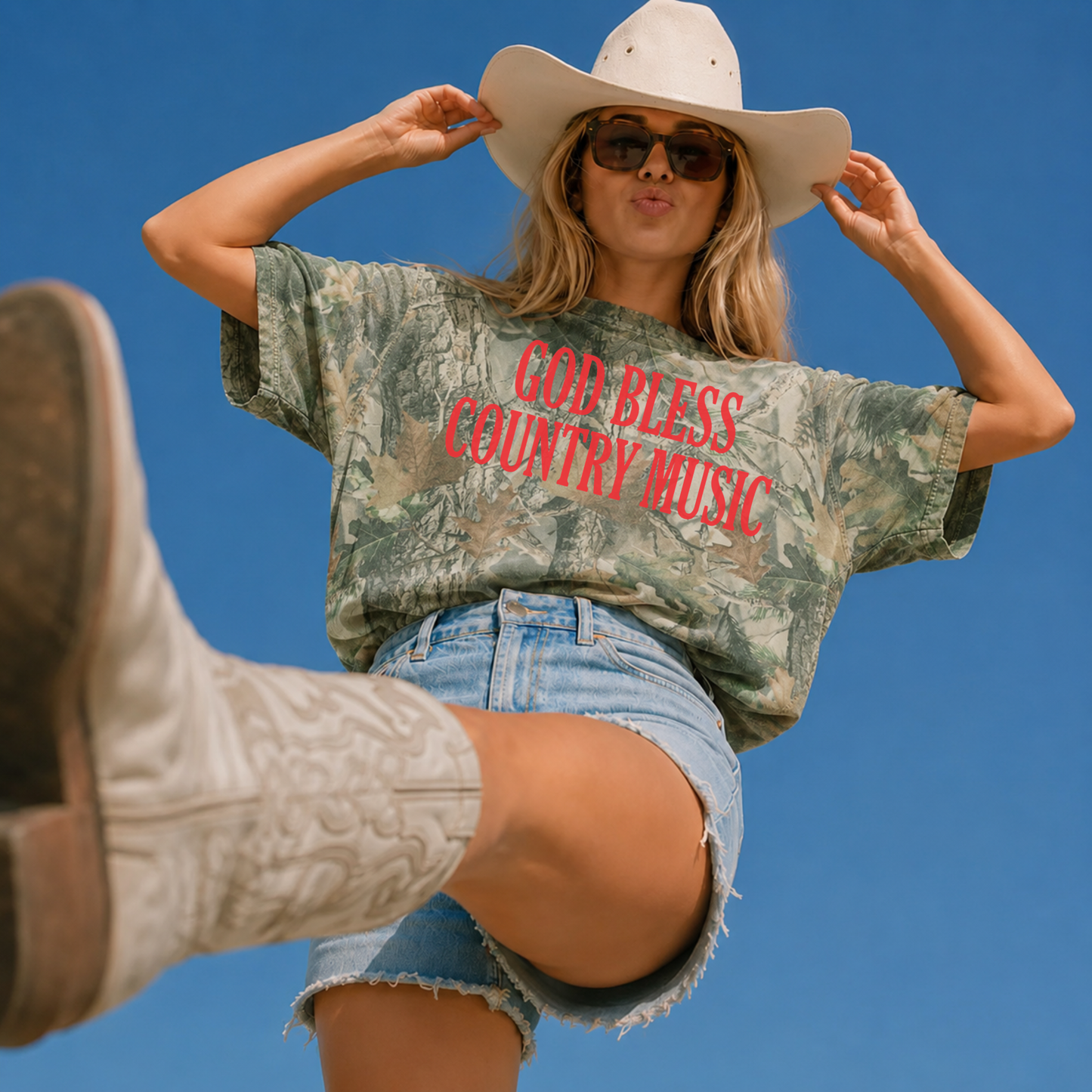 Woman wearing an oversized vintage wood camouflage graphic tee with a bold red "God Bless Country Music" design, styled with denim shorts, white cowboy boots, sunglasses, and a white cowboy hat for a western country concert look.