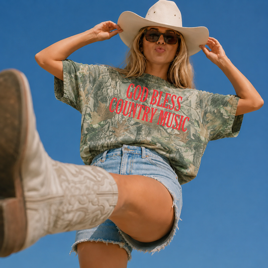 Woman wearing an oversized vintage wood camouflage graphic tee with a bold red "God Bless Country Music" design, styled with denim shorts, white cowboy boots, sunglasses, and a white cowboy hat for a western country concert look.