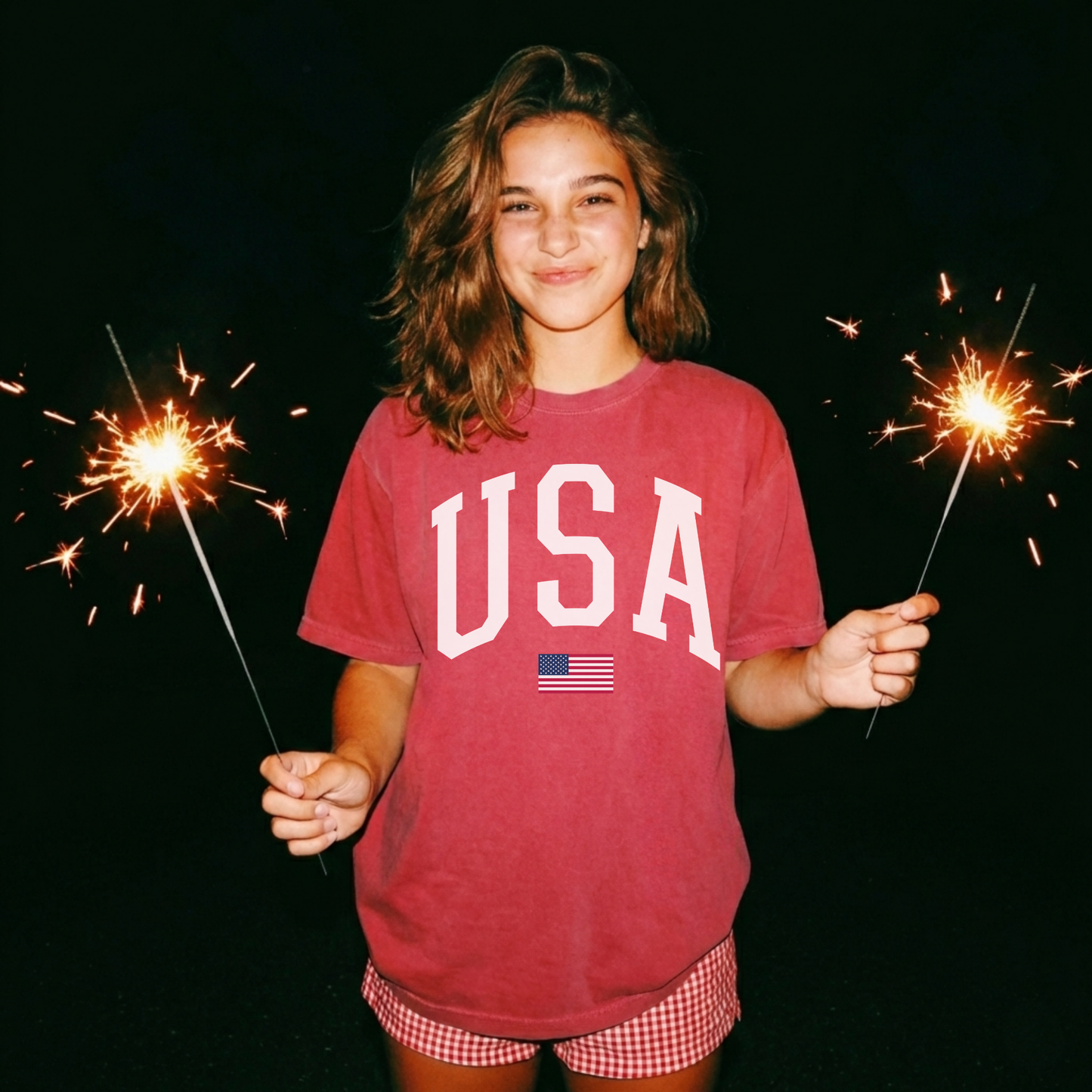 Woman wearing crimson red USA graphic t-shirt with white varsity lettering and small American flag detail, styled for Fourth of July celebration with sparklers.