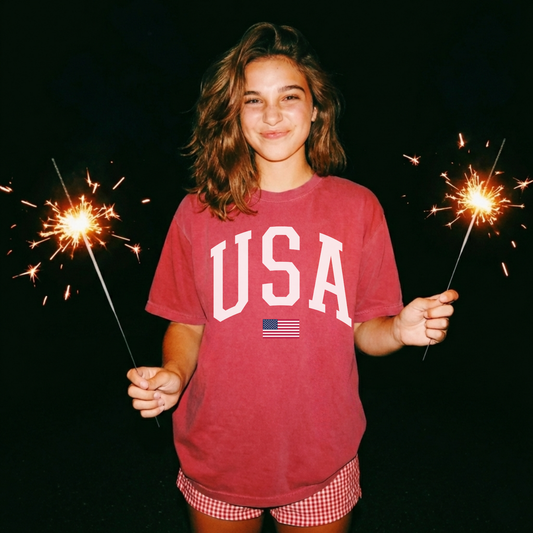 Woman wearing crimson red USA graphic t-shirt with white varsity lettering and small American flag detail, styled for Fourth of July celebration with sparklers.
