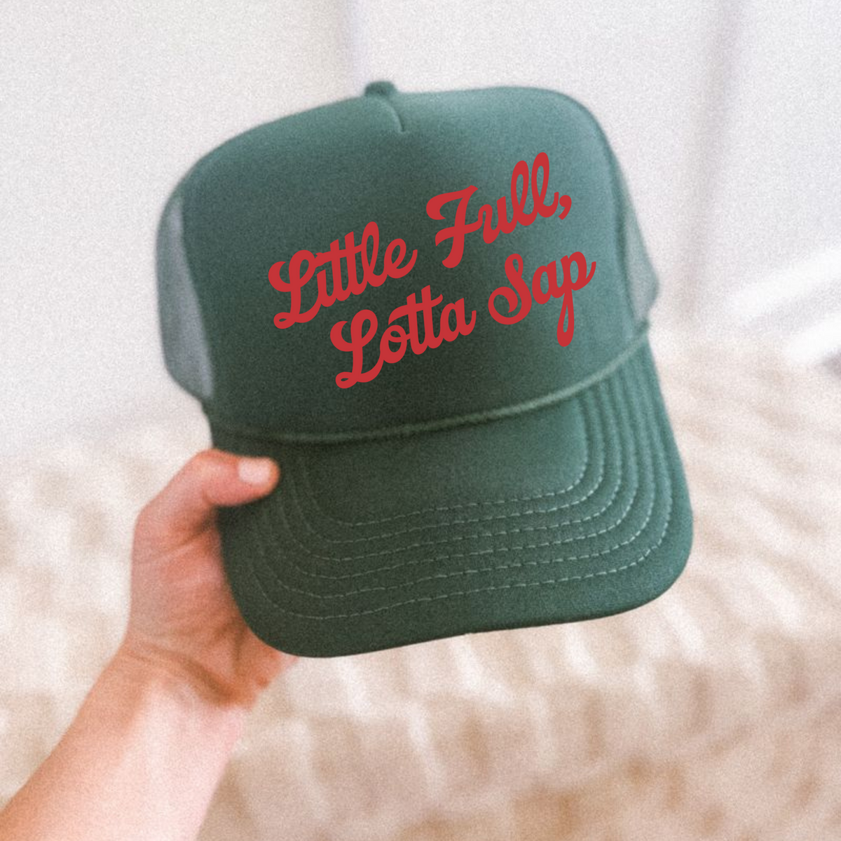Green cap with red text 'Little Full, Lotta Sap' held by a hand on a white background