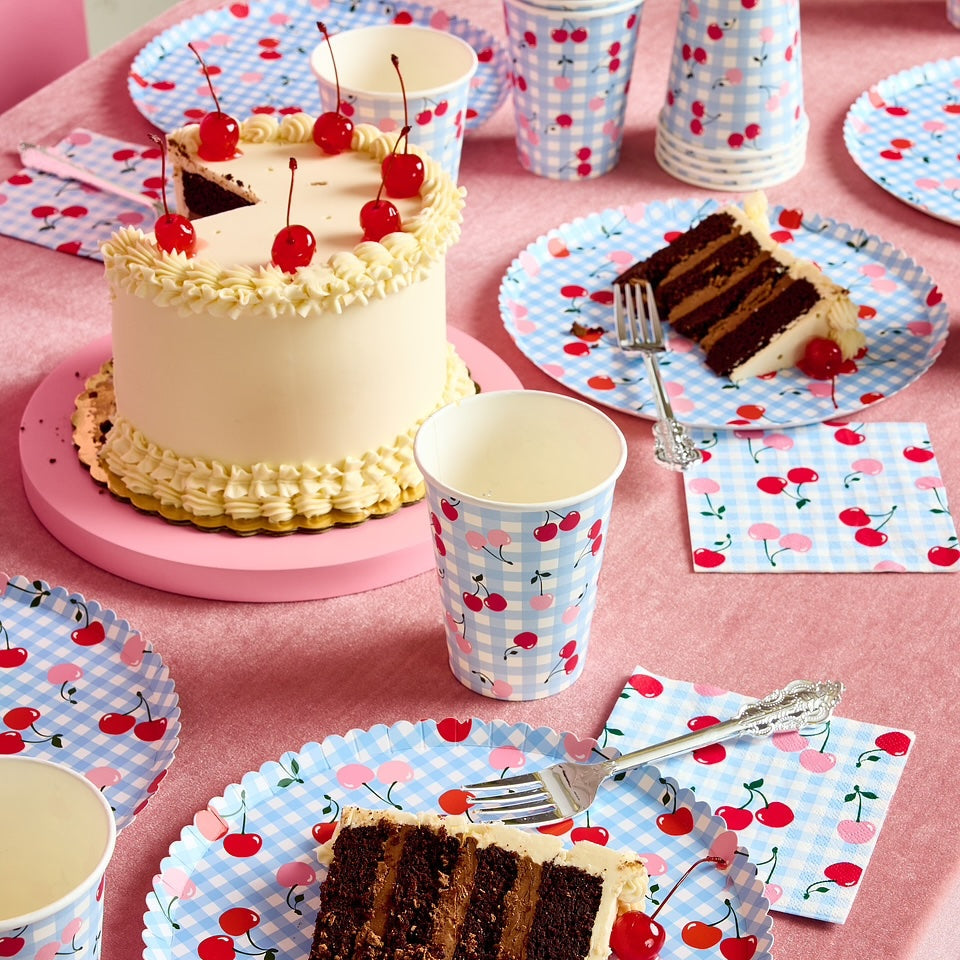 Table setting with cake, cups, and plates featuring cherry designs on a pink tablecloth.