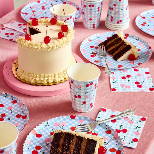 Table setting with cake, cups, and plates featuring cherry designs on a pink tablecloth.