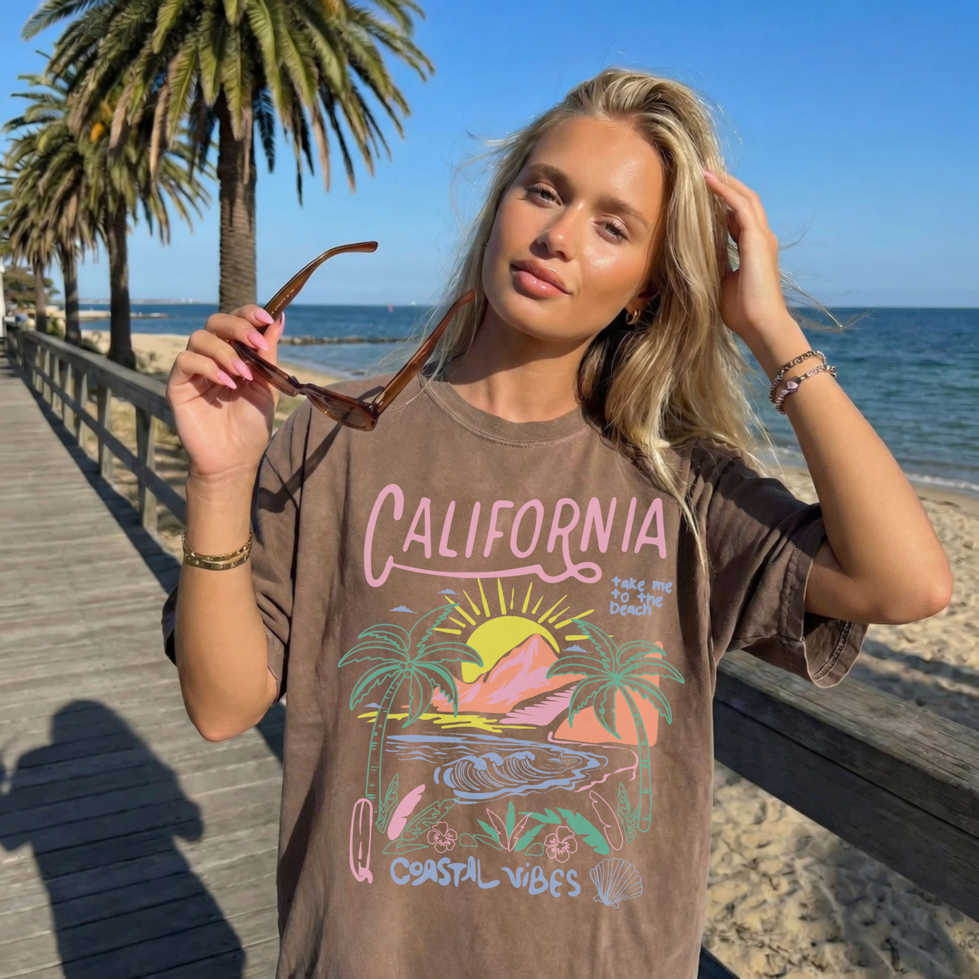 Woman wearing brown California Coastal Vibes graphic tee on beach boardwalk with palm trees