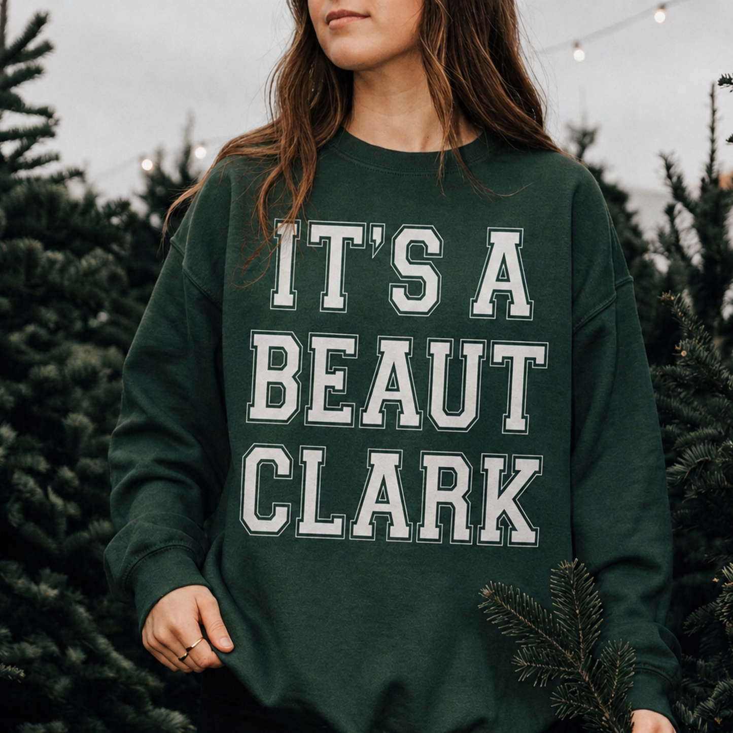 Woman wearing forest green It's a Beaut Clark Christmas sweatshirt with white collegiate lettering at a Christmas tree farm.