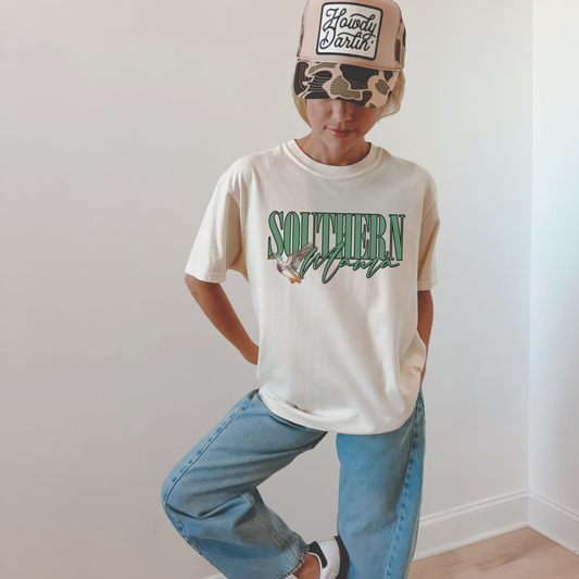 Woman wearing ivory Southern Mama t-shirt with green lettering and flying duck graphic, styled with jeans and a camo hat.