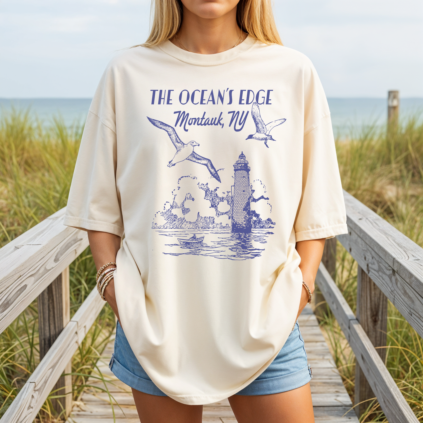 Model wearing true-to-size ivory Comfort Colors Montauk NY tee with blue lighthouse graphic on a beach boardwalk.