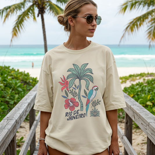 Model wearing a size Large ivory Rio de Janeiro tropical graphic tee on a boardwalk with palm trees and turquoise water.