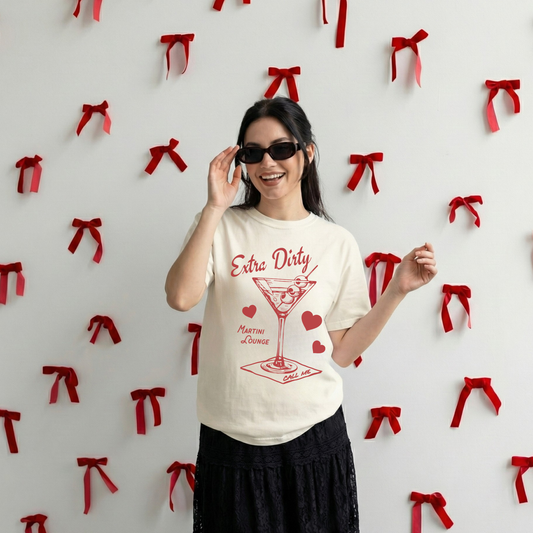 Model wearing ivory Extra Dirty Martini Lounge graphic tee with red martini illustration, standing against wall with red ribbon decorations.