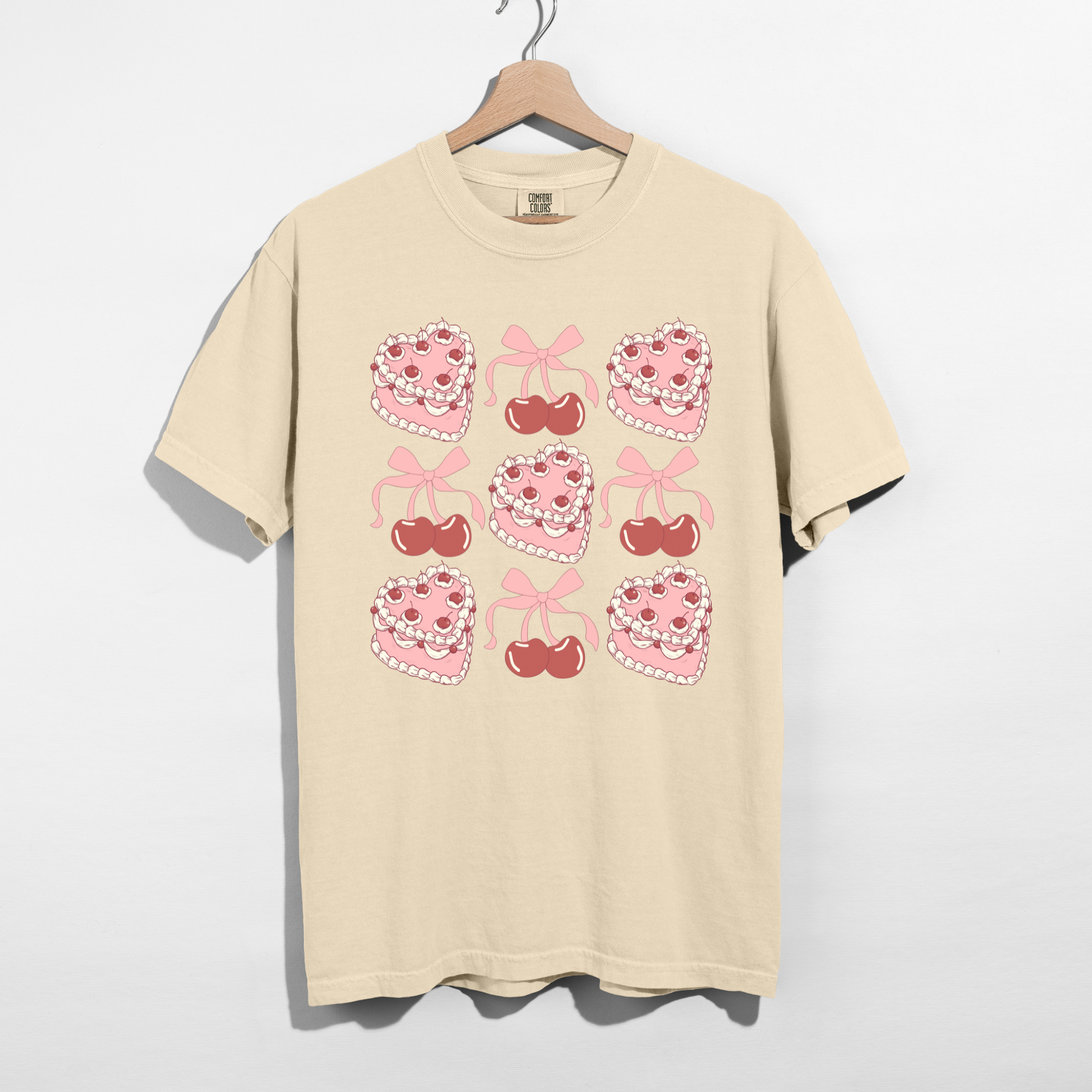 Ivory Sweetheart Cherry Cake Graphic Tee featuring retro pink heart cakes, cherries, and bows displayed on a hanger.