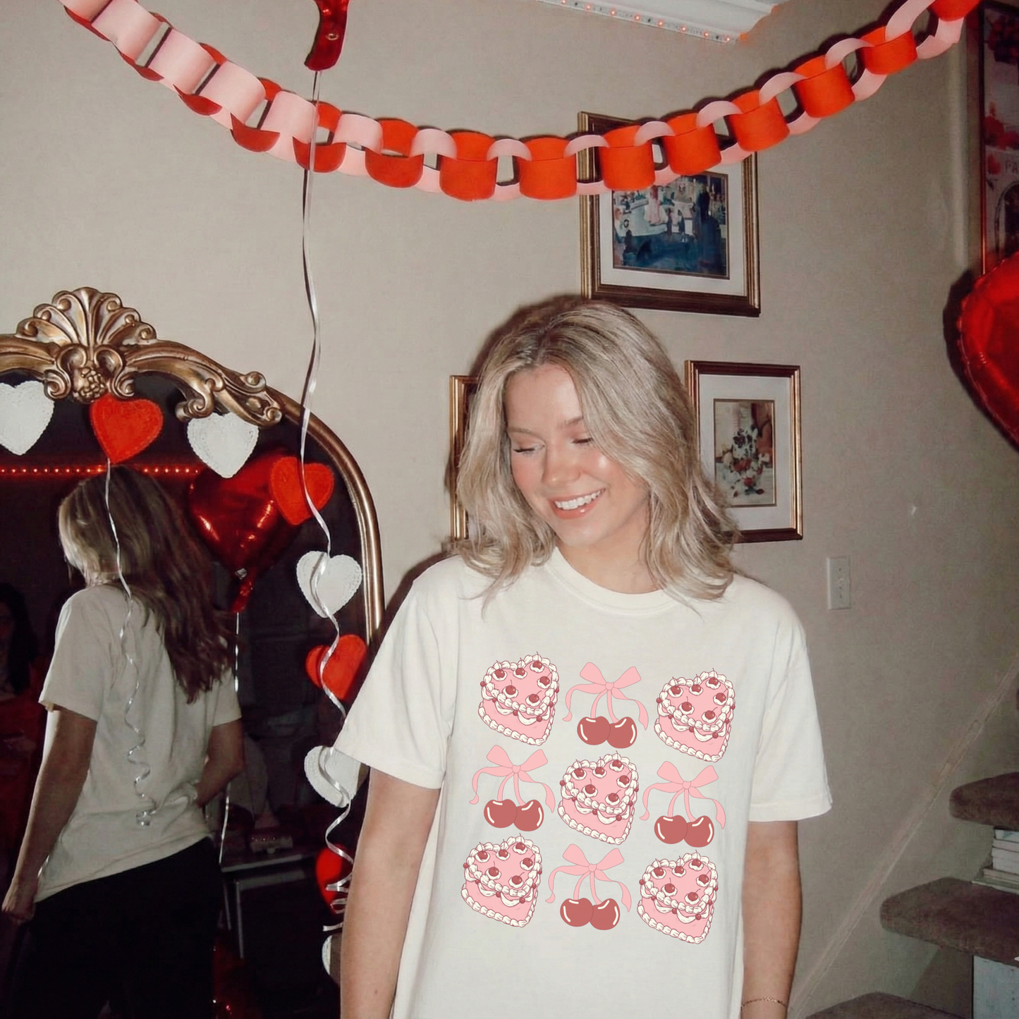 Woman wearing an ivory Sweetheart Cherry Cake Graphic Tee standing in a Valentine-themed room with heart decorations.