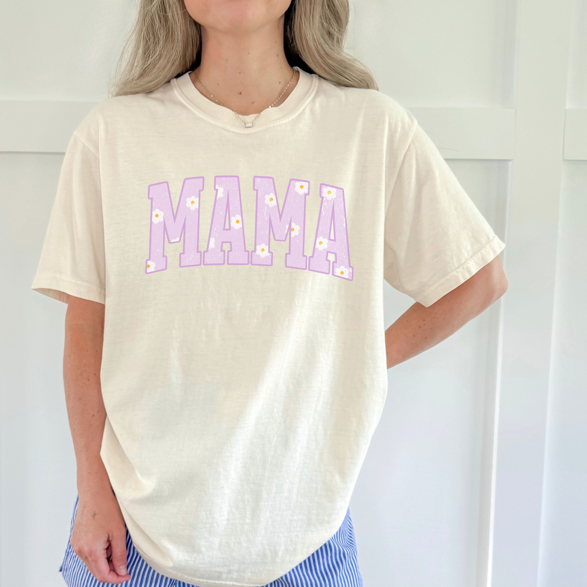 Woman wearing an ivory Comfort Colors mama graphic t-shirt with pastel purple daisy lettering, relaxed true to size fit