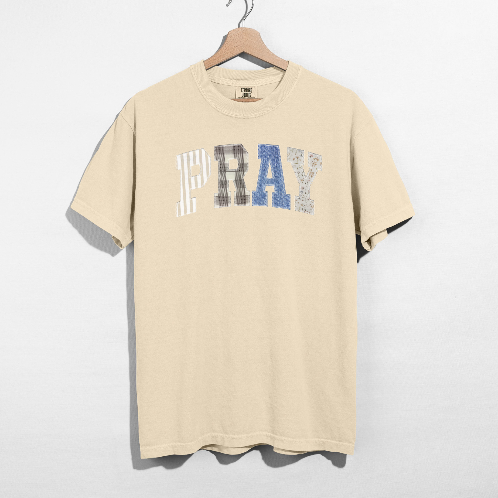 Beige t-shirt with 'PRAY' text on a hanger against a white background