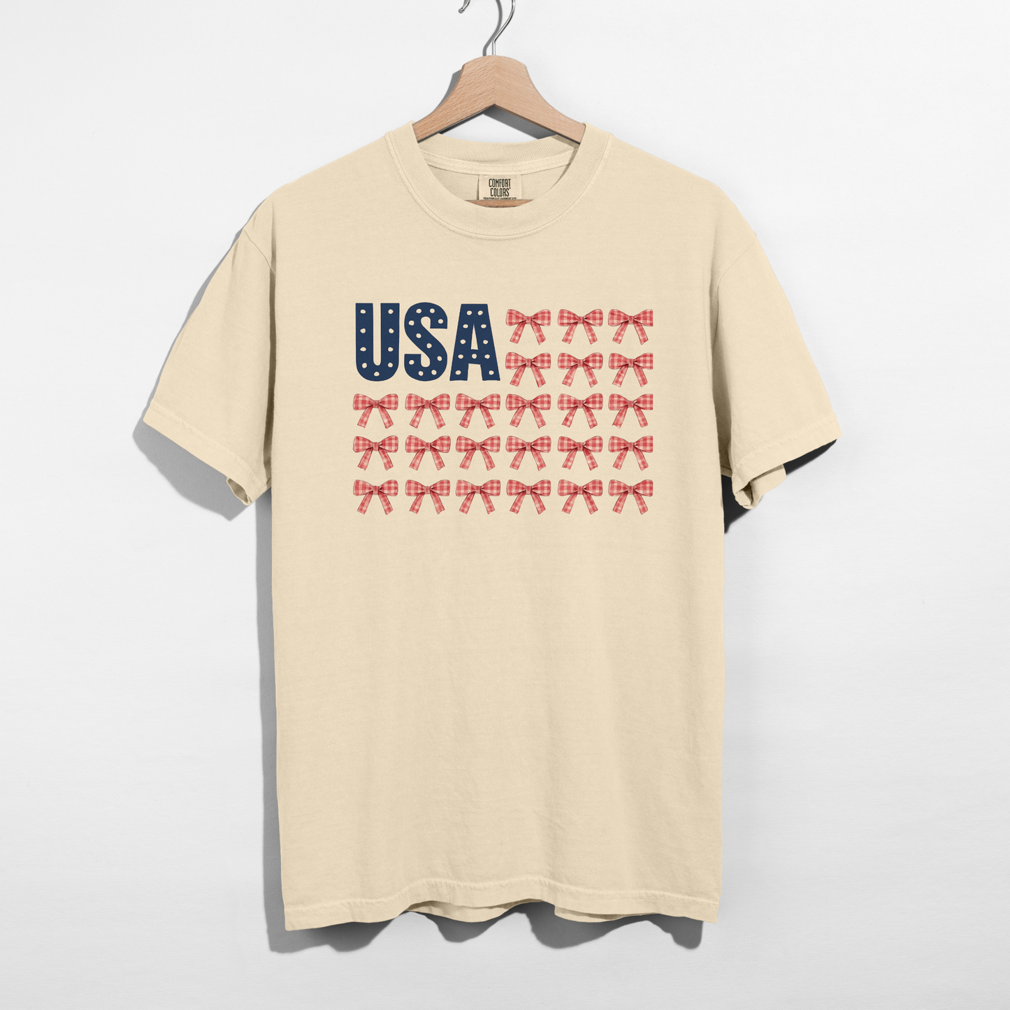 Beige t-shirt with 'USA' and bow design on a hanger against a white background