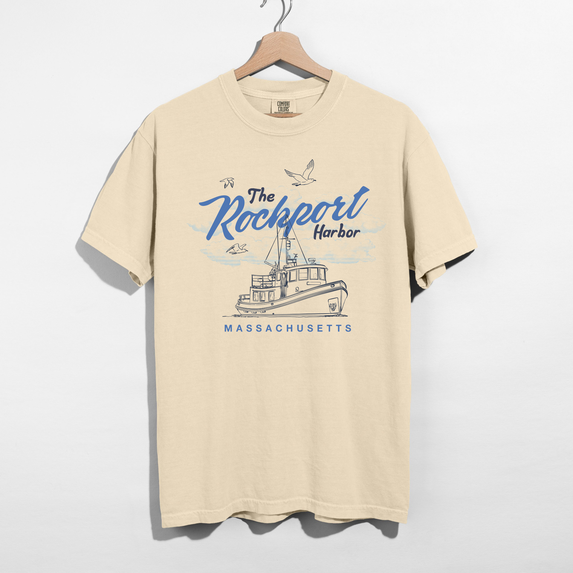 Beige t-shirt with 'The Rockport Harbor, Massachusetts' graphic on a hanger against a white background