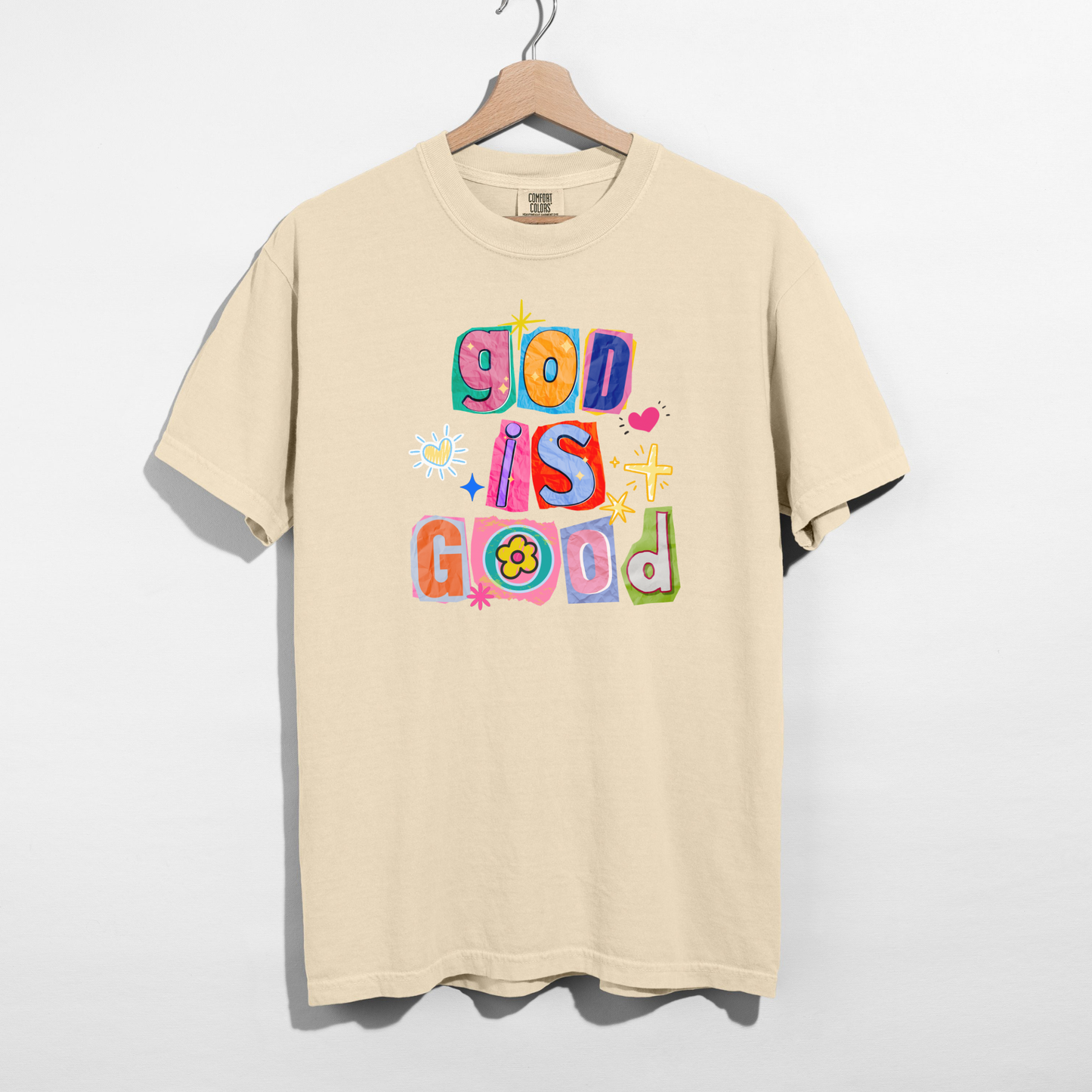 Beige t-shirt with colorful text 'God is Good' on a hanger against a white background