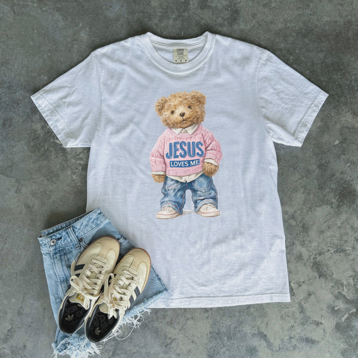 White t-shirt with teddy bear graphic and text, paired with jeans and sneakers on a gray surface
