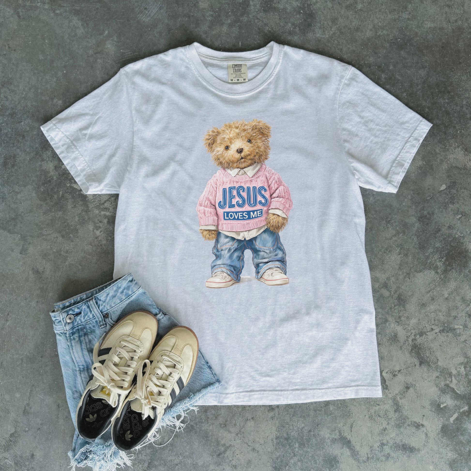 White t-shirt with teddy bear graphic and text, paired with jeans and sneakers on a gray surface