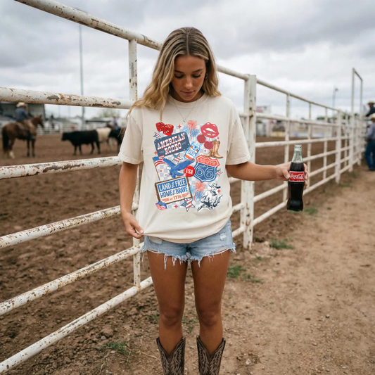 Ivory American Rodeo graphic t-shirt styled with denim shorts and cowboy boots at a rodeo arena featuring patriotic western collage design.