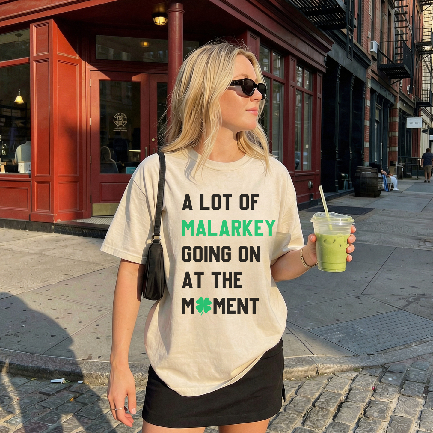 Woman wearing ivory A Lot of Malarkey Going On at the Moment St Patrick’s Day graphic tee holding green drink