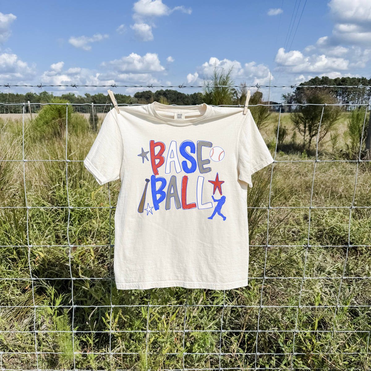Ivory baseball graphic t-shirt with red white and blue retro lettering hanging on wire fence in sunny rural field setting.