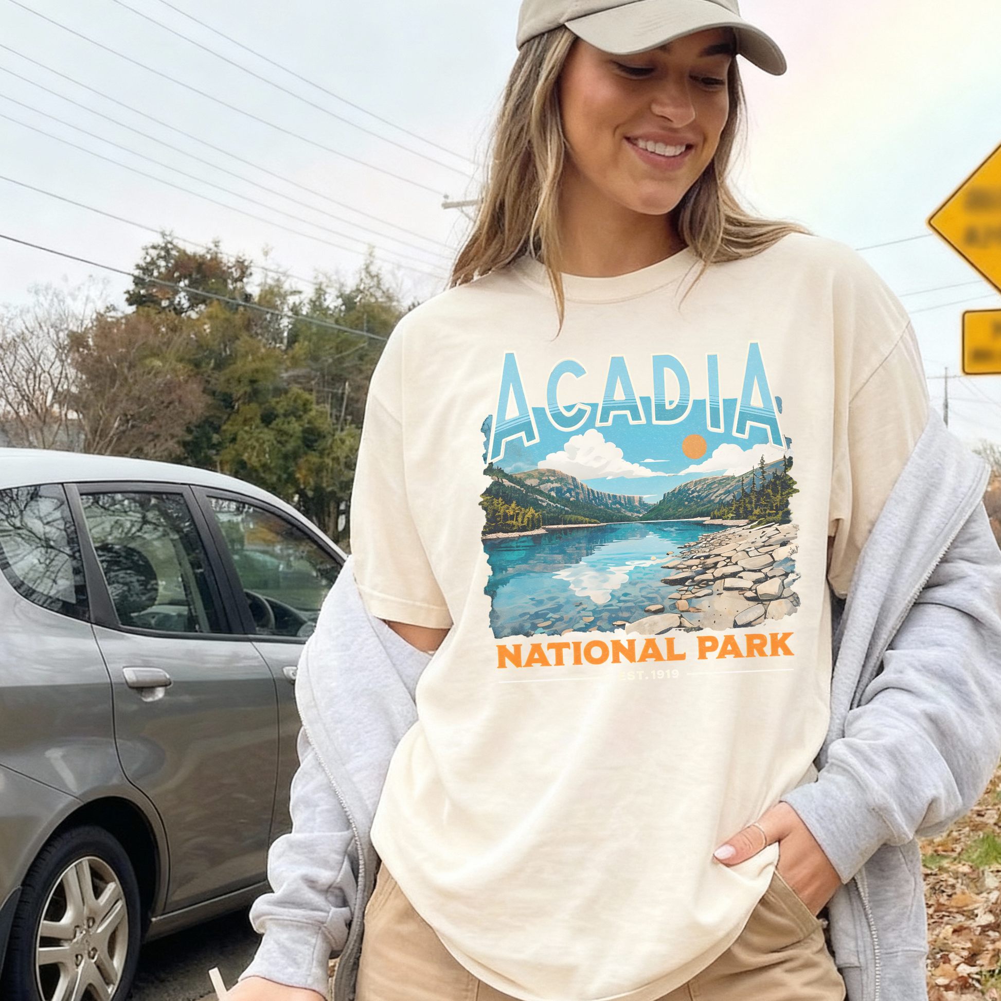 Woman wearing ivory Acadia National Park retro travel t-shirt with mountain lake design layered with grey sweatshirt and cap.