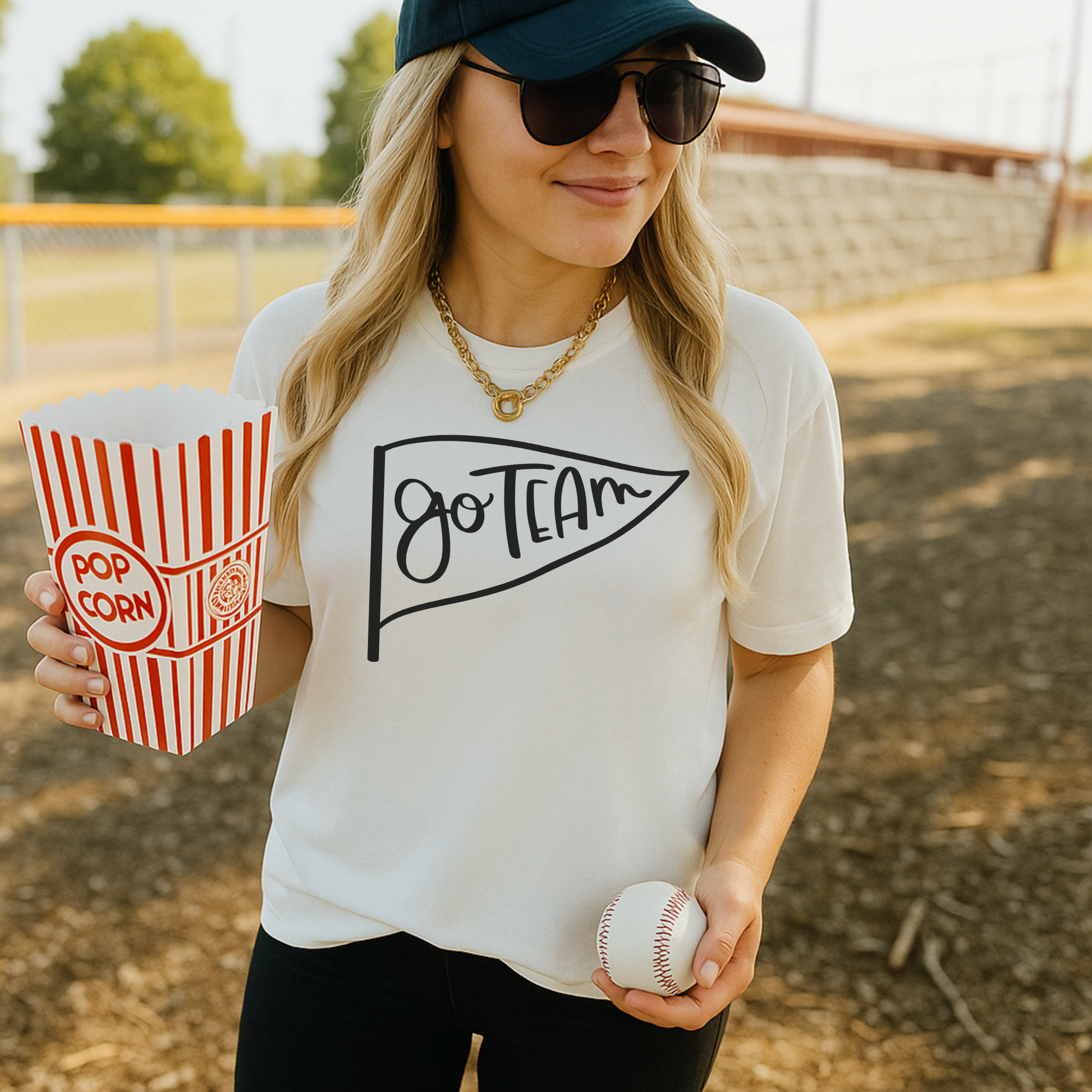 Woman at a baseball game holding popcorn and a ball, wearing a 'Go Team' shirt.