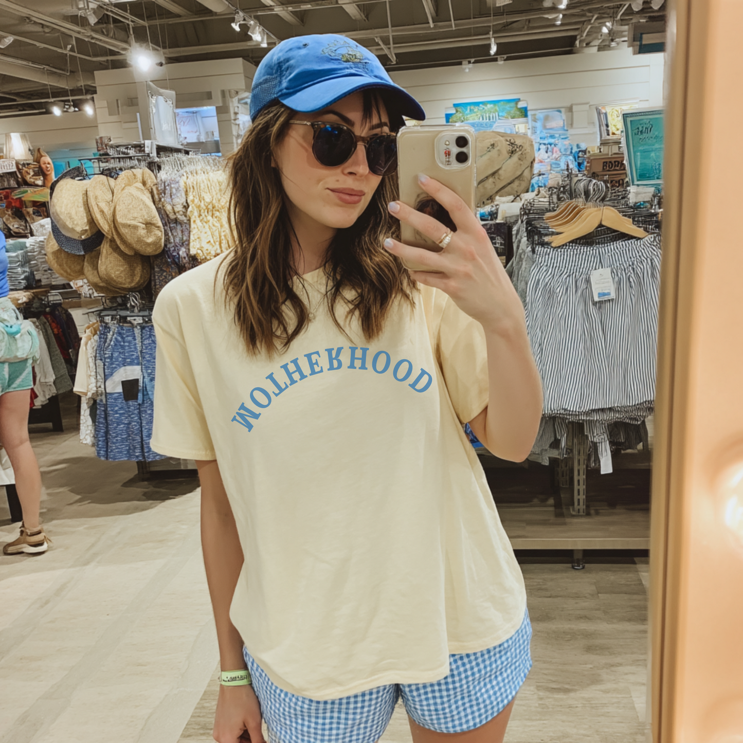 A person wearing a beige t-shirt with the text 'MOTHERHOOD' in bold blue letters, paired with blue and white checkered shorts and a blue baseball cap, taking a mirror selfie in a retail environment.