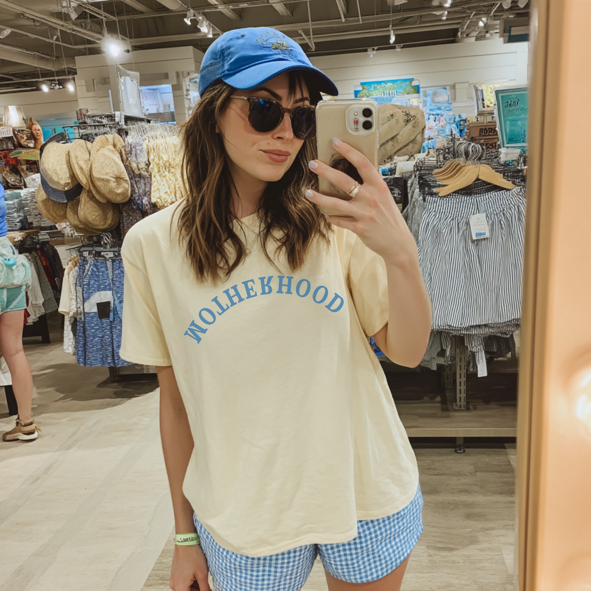 A person wearing a beige t-shirt with the text 'MOTHERHOOD' in bold blue letters, paired with blue and white checkered shorts and a blue baseball cap, taking a mirror selfie in a retail environment.