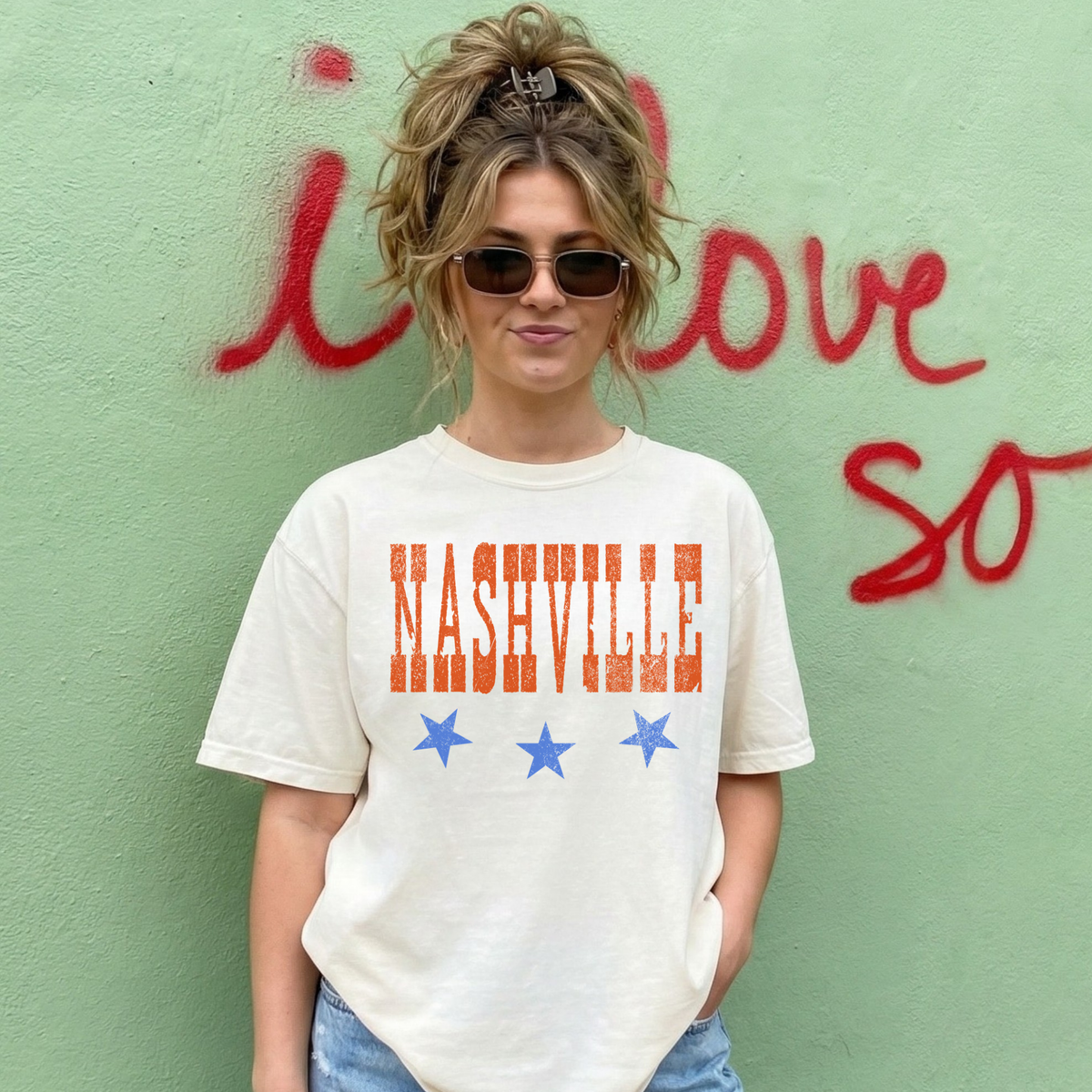 Woman wearing ivory Nashville graphic tee with orange text and blue stars casual outfit against green wall