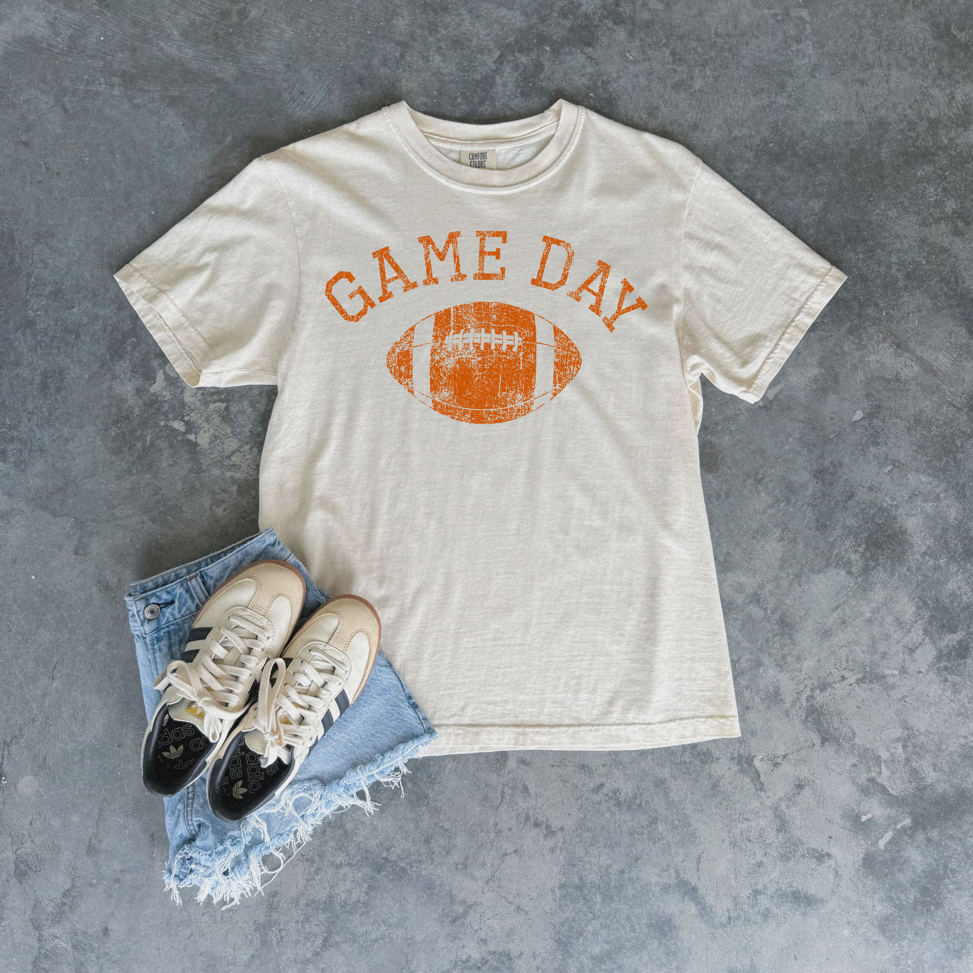 An off-white t-shirt with a distressed orange 'GAME DAY' football graphic, styled in a flat lay with light blue frayed denim shorts and white sneakers on a textured gray background.
