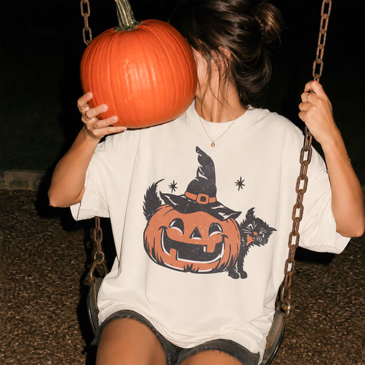 Woman wearing an ivory vintage Halloween graphic tee featuring a smiling jack-o'-lantern, witch hat, and black cat while holding a pumpkin on a swing at night.
