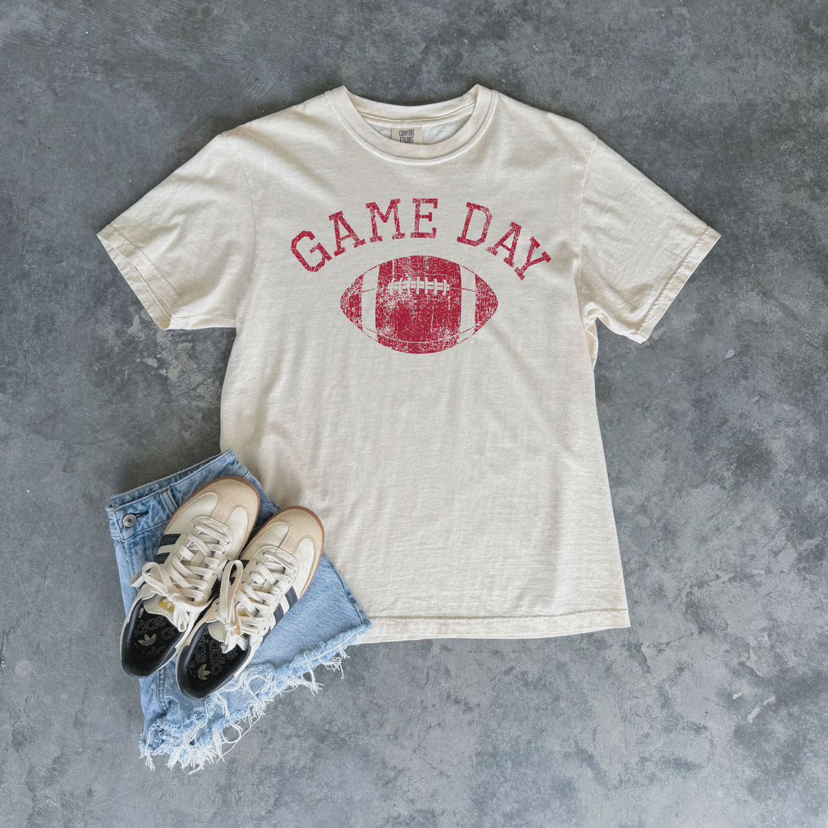 An off-white crewneck t-shirt with a distressed red 'GAME DAY' football graphic, styled in a flat lay with light blue denim shorts and beige/black sneakers on a textured gray background.
