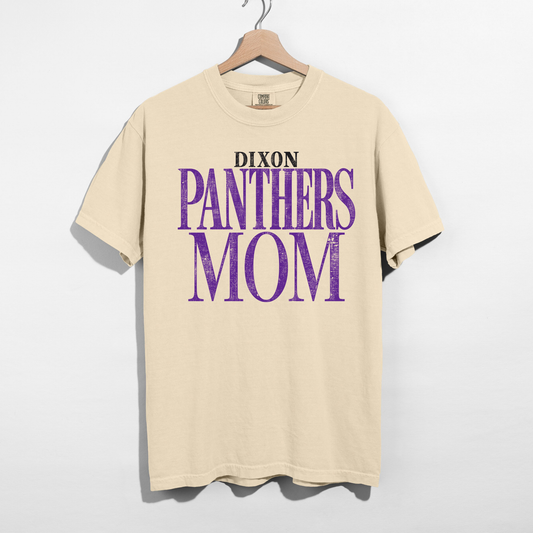 A beige short-sleeve t-shirt hanging on a wooden hanger against a light gray wall. The shirt features a graphic print on the front that reads 'DIXON PANTHERS MOM' in black and distressed purple lettering.