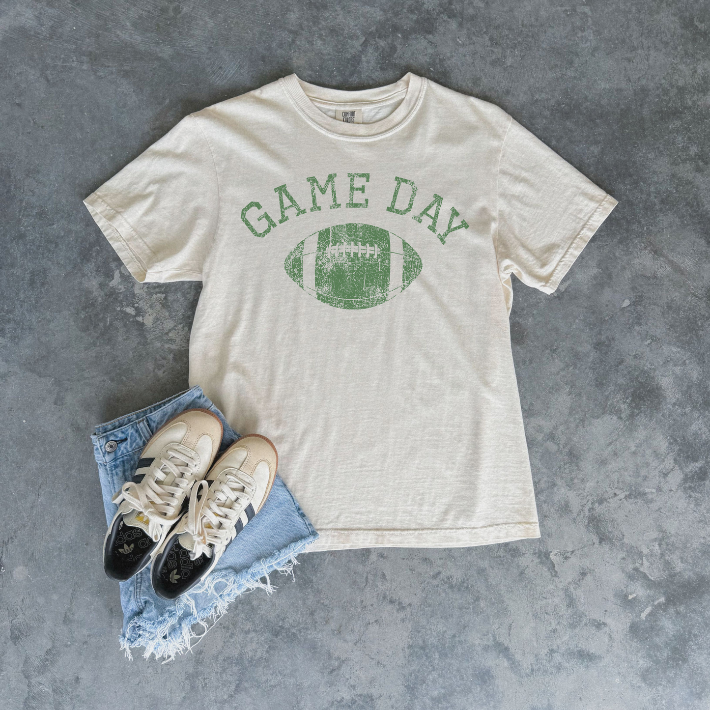 An off-white crewneck t-shirt with a green distressed 'GAME DAY' football graphic, styled in a flat lay with light blue denim shorts and white sneakers on a textured gray background.