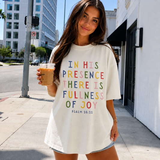 Woman wearing an ivory Christian graphic t-shirt featuring the colorful Bible verse In His Presence There Is Fullness of Joy Psalm 16:11 while holding an iced coffee.