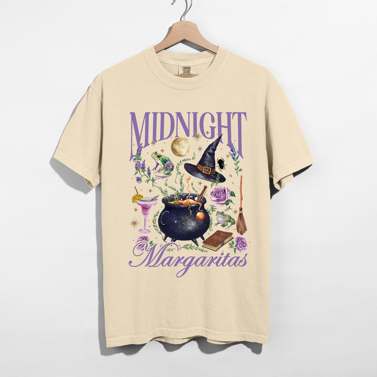 Ivory Midnight Margaritas graphic t-shirt on a wooden hanger featuring purple lettering, magical cauldron, witch hat, margarita cocktail, full moon, frogs, and purple flowers.