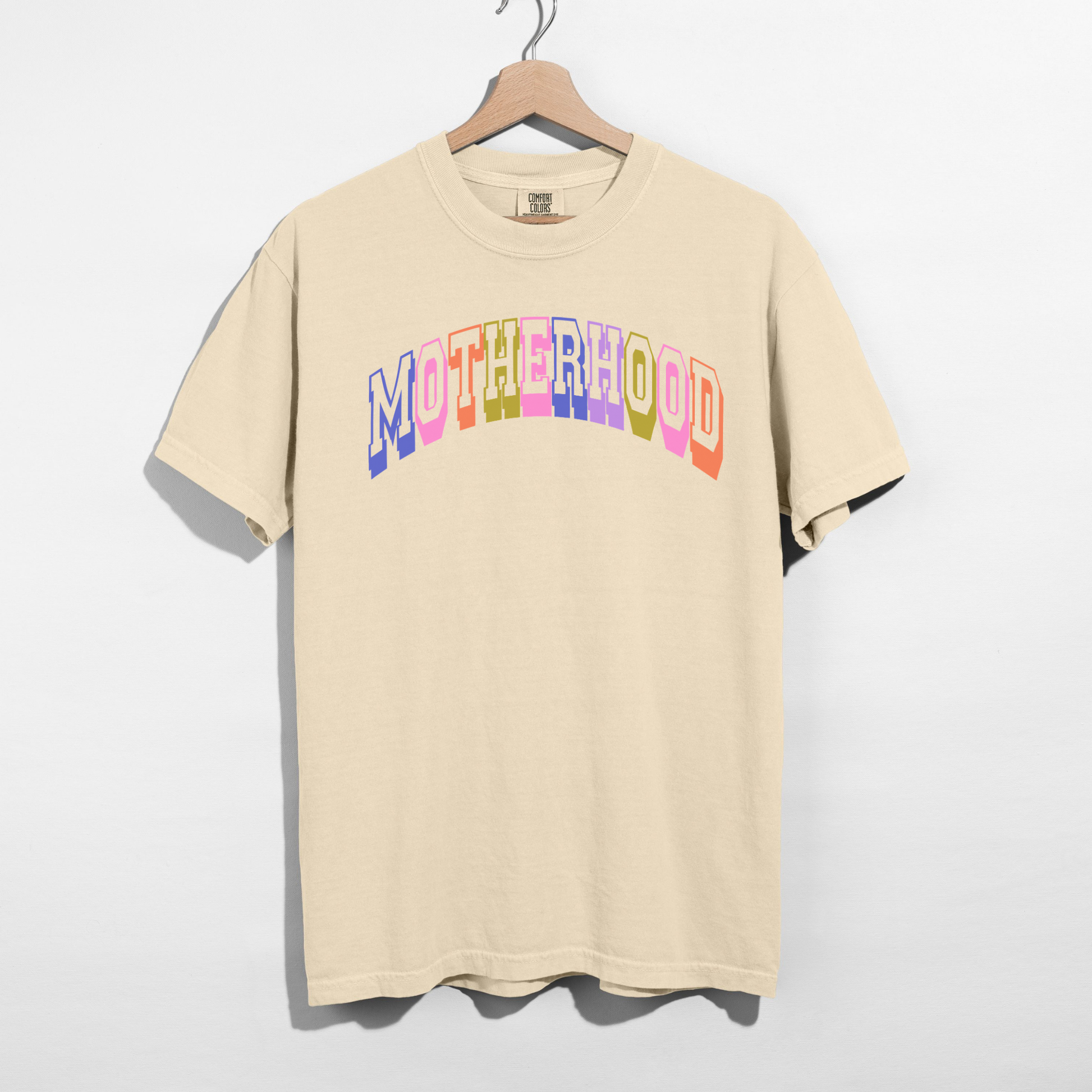 Ivory Comfort Colors 1717 retro motherhood graphic tee with pastel collegiate lettering hanging on wooden hanger.