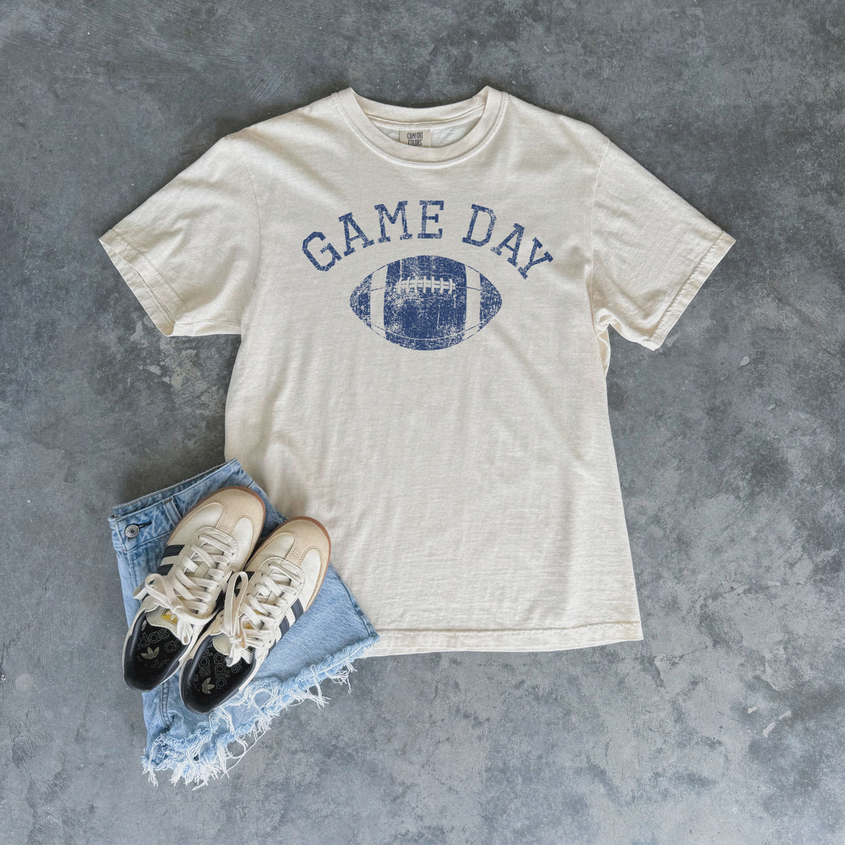 An off-white t-shirt with a blue distressed 'GAME DAY' football graphic, styled in a flat lay with light-wash denim shorts and sneakers on a textured gray background.