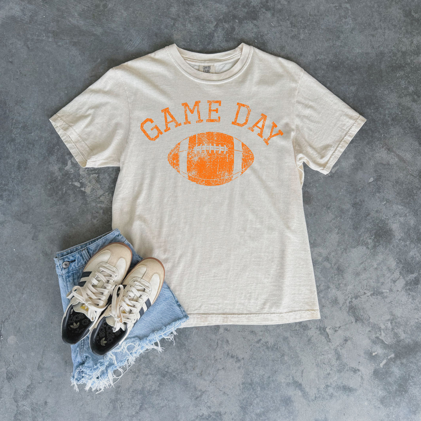 An off-white t-shirt with an orange distressed 'GAME DAY' football graphic, styled in a flat lay with light blue frayed denim shorts and white sneakers on a textured gray background.