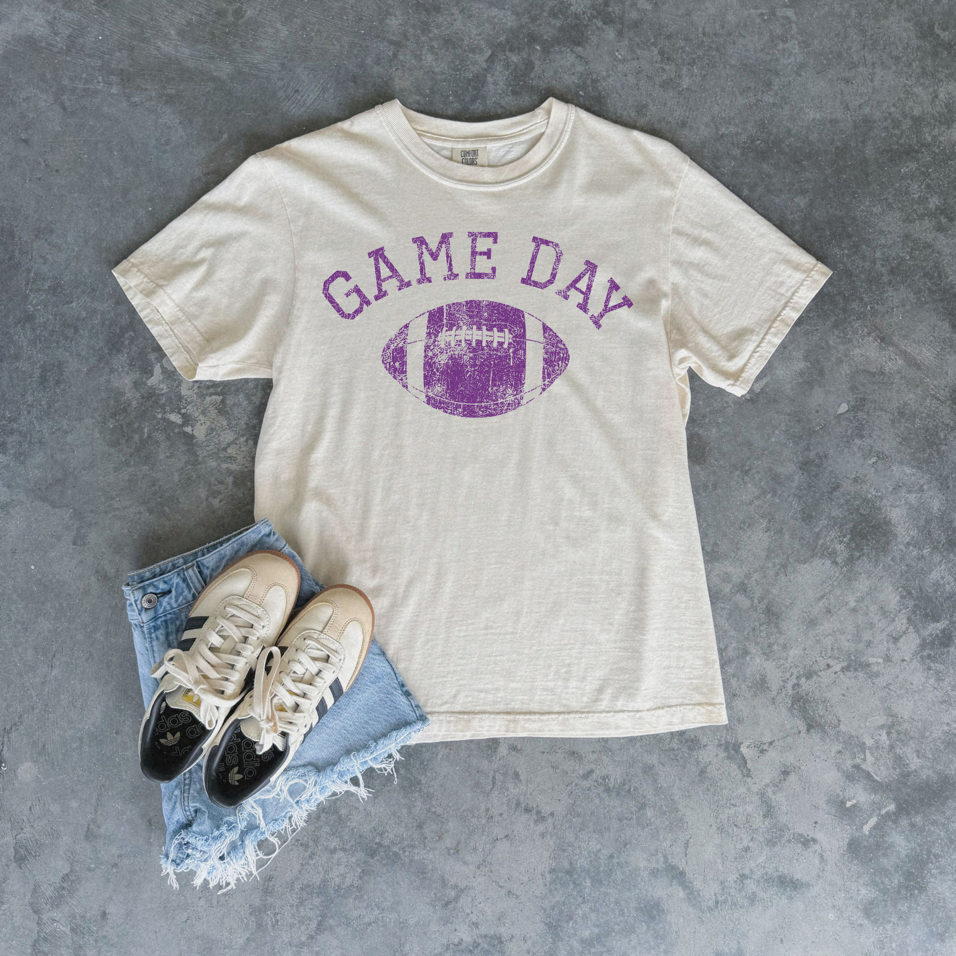 An off-white t-shirt with a purple distressed 'GAME DAY' football graphic, styled in a flat lay with light blue denim shorts and beige sneakers on a textured gray background.