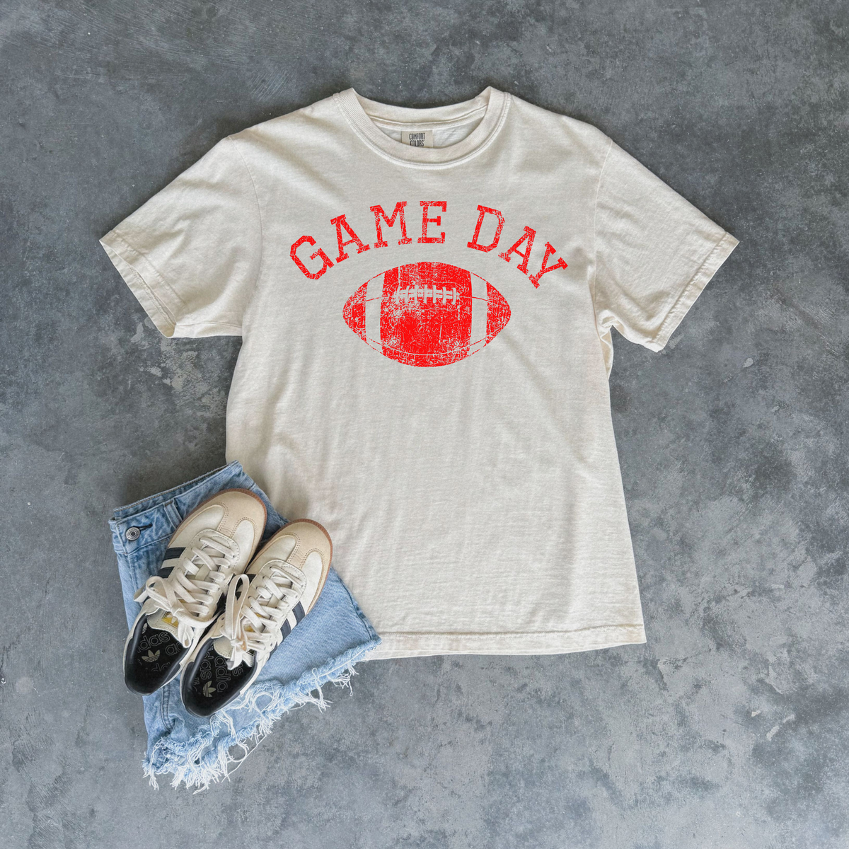 An off-white t-shirt with a red distressed 'GAME DAY' football graphic, styled in a flat lay with light blue frayed denim shorts and beige/black sneakers on a textured gray background.