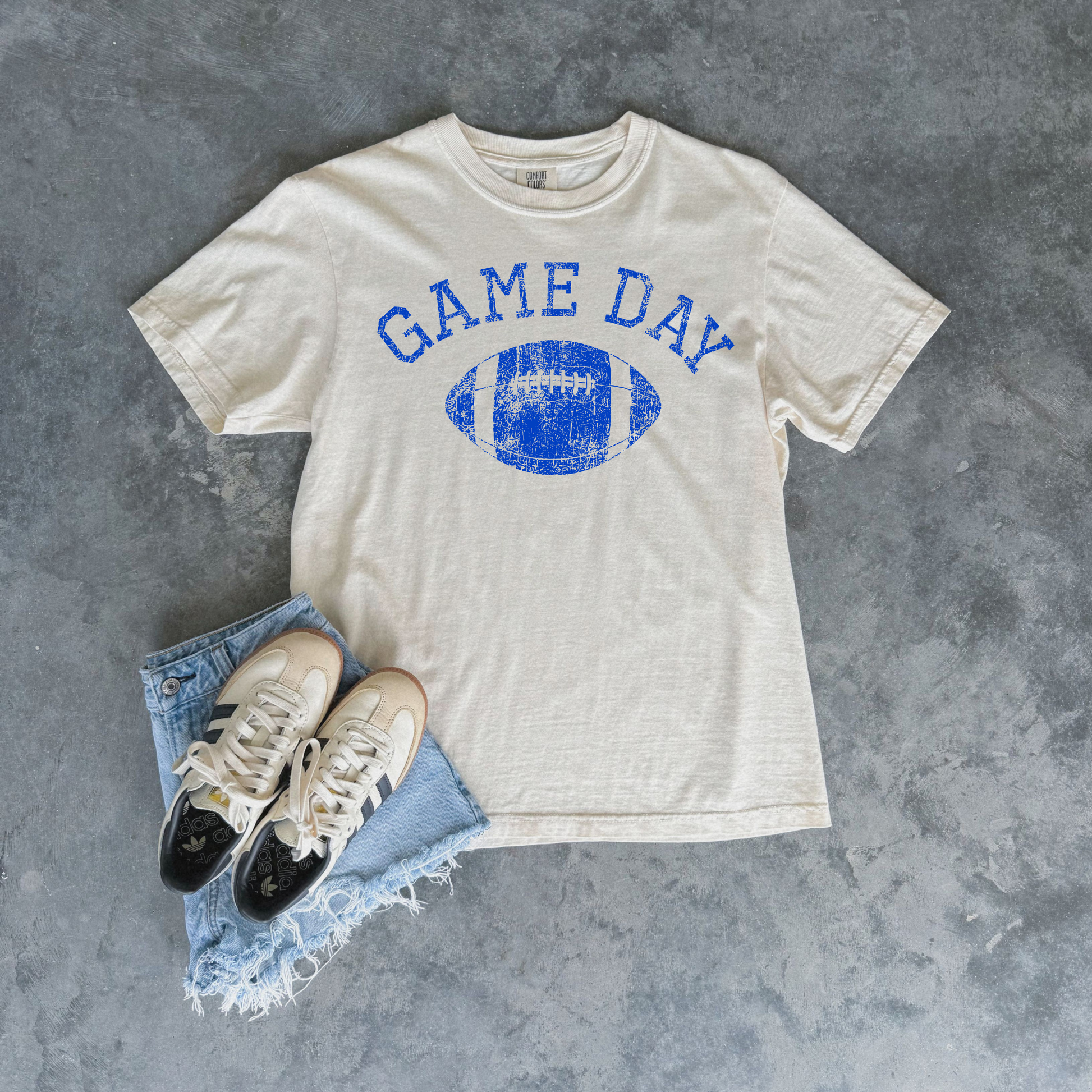 An off-white t-shirt with a blue distressed 'GAME DAY' football graphic, styled in a flat lay with light blue denim shorts and beige sneakers on a textured gray background.