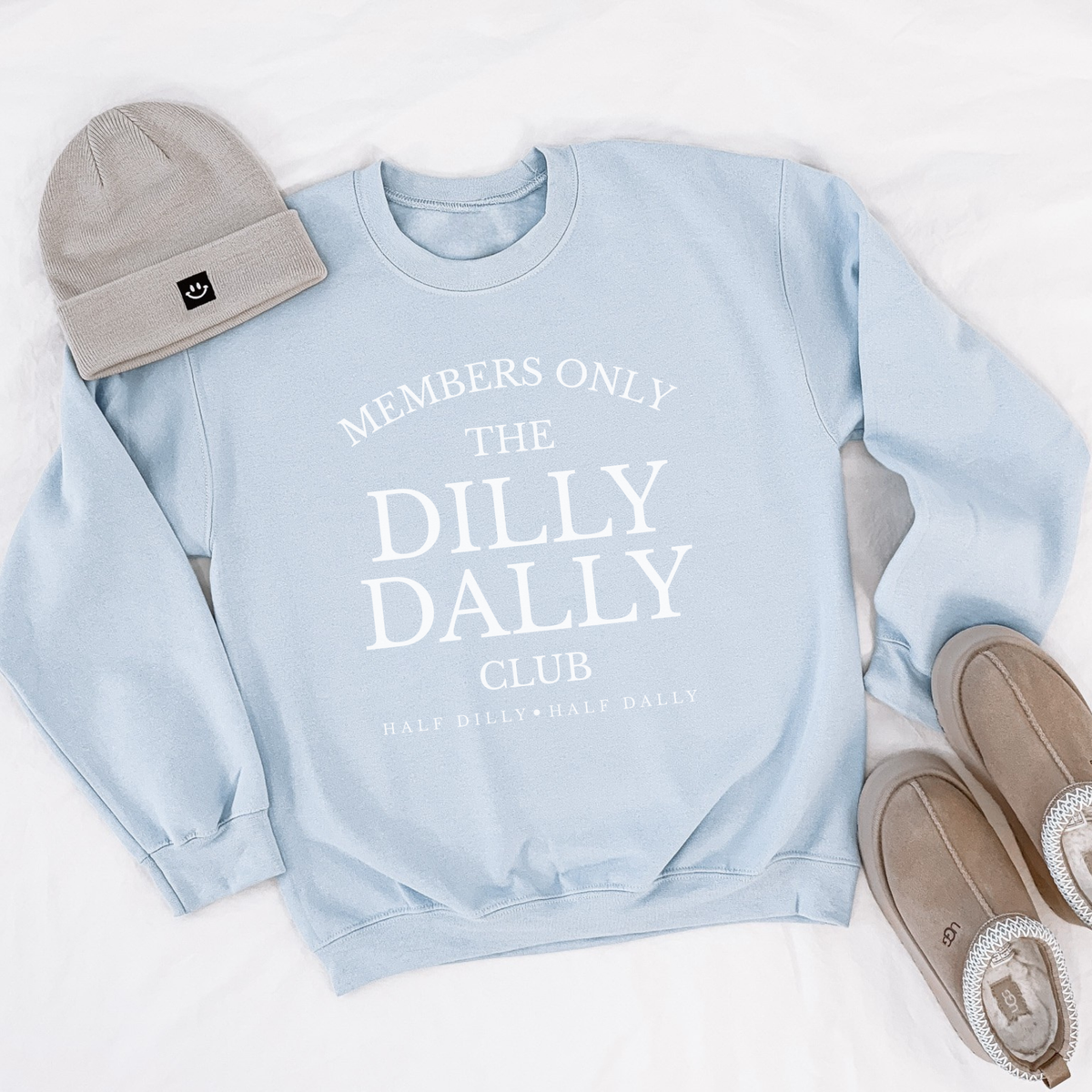 Light blue “Dilly Dally Club” crewneck sweatshirt styled with a beige beanie and slippers.