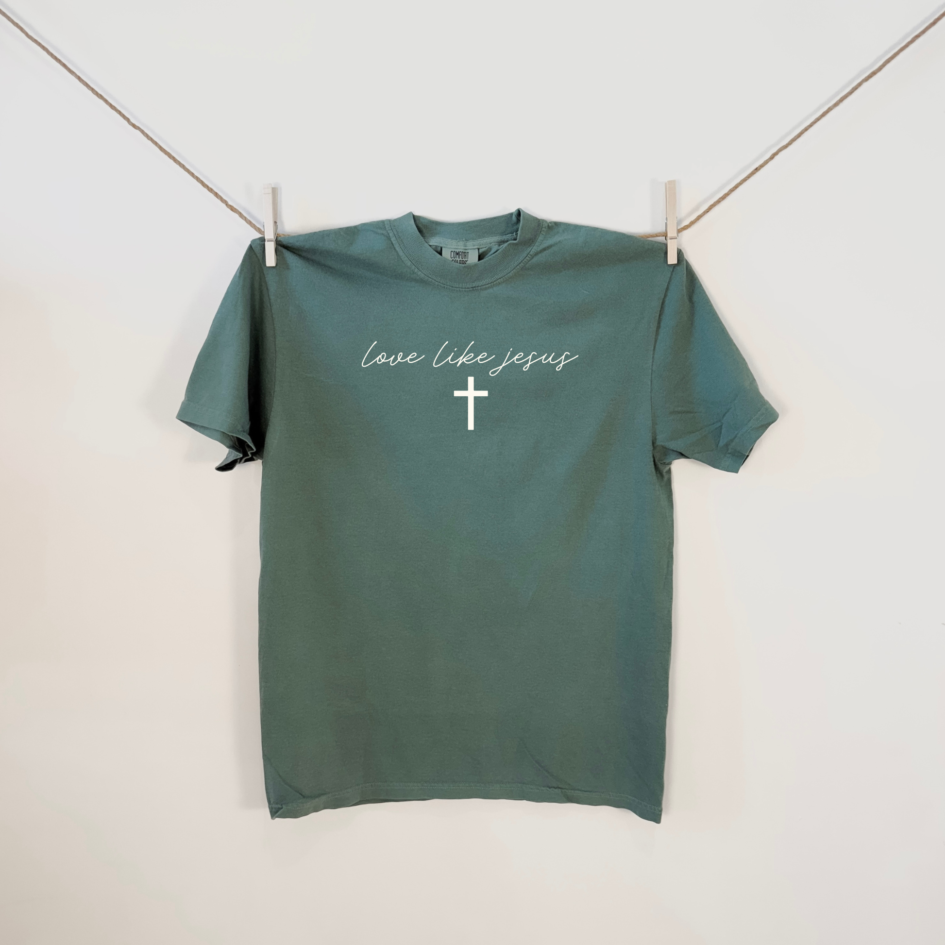 Light Green Christian t-shirt hanging on line with “Love Like Jesus” script and cross graphic.