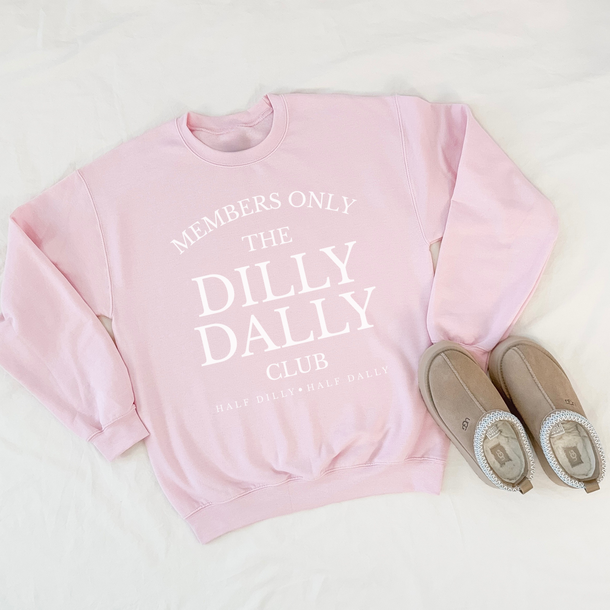 Light pink “Dilly Dally Club” sweatshirt laid flat with slippers for cozy styling.