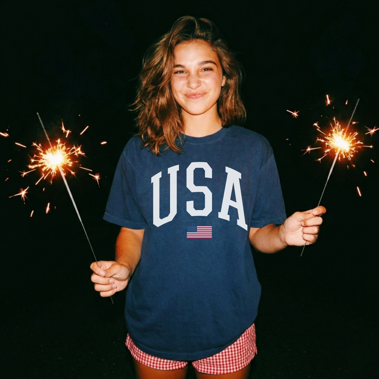 Woman wearing navy USA patriotic graphic tee with white collegiate font and American flag detail while holding sparklers at night.